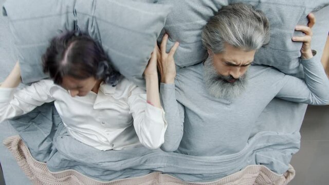 Irritated couple mature man and young woman are fighting expressing anger lying in bed together covering heads with pillows avoiding communication