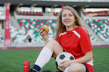 Portrait of young female football player sitting on soccer field with apple after the training