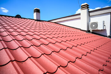 House roof. Red metal roof tiles. Roof made of metal sheets with cloudy sky in background. Air conditioner on a house wall. 