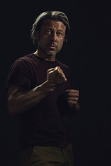 Dark portrait of a middle-aged man with blond hair and a gray stubble in a dark brown t-shirt with clenched fists.
