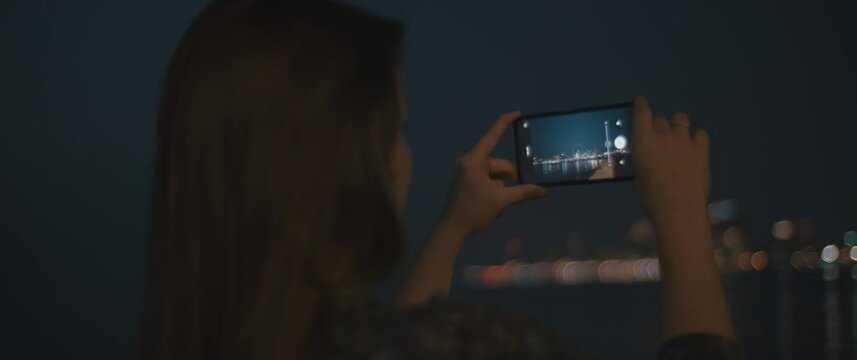 Close up of a young woman using a smartphone to take pictures of a night cityscape. Slow motion, close up. 