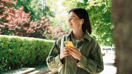 Young Woman Texting on Her Smartphone While Walking in the City Park, Brunette Girl Using Cell Phone and Earphones Looking Away