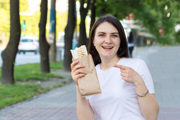 A beautiful brunette girl keeps a shawarma in the street. Food on the way on the way to work. Fast food cafe advertising.