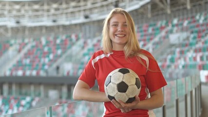 Portrait of young caucasian woman in red football strip holding ball at big soccer stadium, smiling to camera - Powered by Adobe