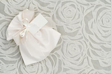 Beautiful floral pattern background with wedding favor, tag and bow mock-up. Abstract design with ribbon and ivory pouch on left side. Blank horizontal image,  flat lay.
