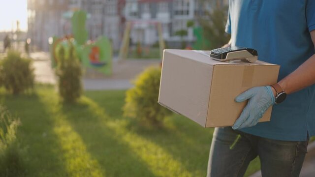 A male deliveryman in gloves brings a box and gives the terminal to pay for the purchase. Carry a box with an NFC credit card payment terminal or mobile phone. Contactless payment