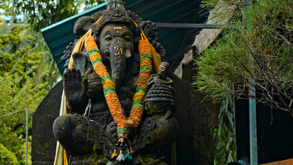 An old stone statue of Ganesha covered with moss with a wreath of yellow flowers on his neck standing on the street