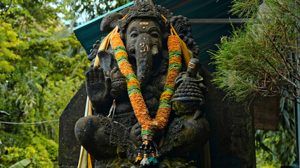 An old stone statue of Ganesha covered with moss with a wreath of yellow flowers on his neck standing on the street