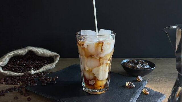 Pouring milk into a glass with iced coffee over black background. Cold refreshment summer coffee latte drink.