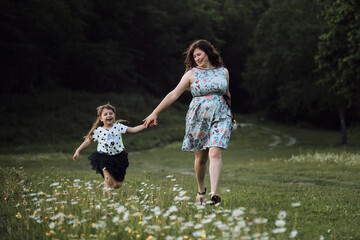 Fototapeta premium Have fun with child in nature, the joy of motherhood. Young beautiful European mother in blue dress runs through chamomile field with her little daughter and laughs.