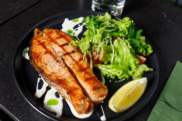 One piece of baked salmon grilled pepper lemon and salt on a black plate with lettuce leaves on a stone background