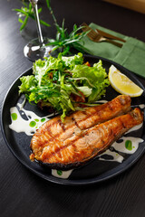 One piece of baked salmon grilled pepper lemon and salt on a black plate with lettuce leaves on a stone background