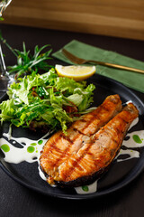 One piece of baked salmon grilled pepper lemon and salt on a black plate with lettuce leaves on a stone background
