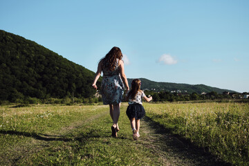 Have fun with child in nature, the joy of motherhood. Young beautiful European mother in blue dress runs through chamomile field with her little daughter and laughs. Rear view.