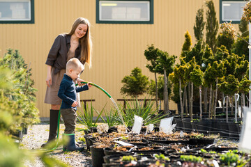 Little son helps mom to water the pots of plants in garden center.