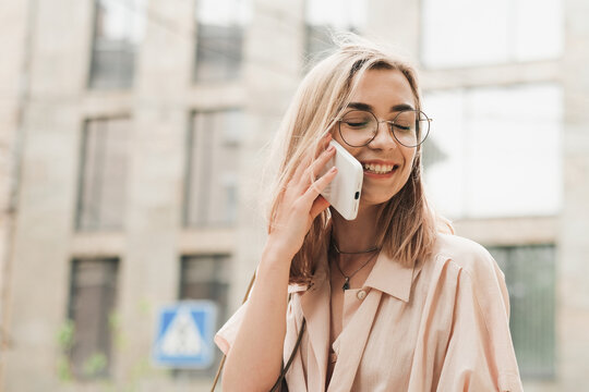 Cheerful Elegant Woman in Glasses Using Smartphone and Smiling with Closed Eyes, Stylish Blonde Girl Outdoors Standing on the Street Having Conversation Through Mobile Phone