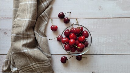 Sweet red cherries in glass bowl, on white wooden old table and brown napkin next to the bowl, selective focus. Not edited photo, raw image. Copy space available. Under the sun. Unedited.