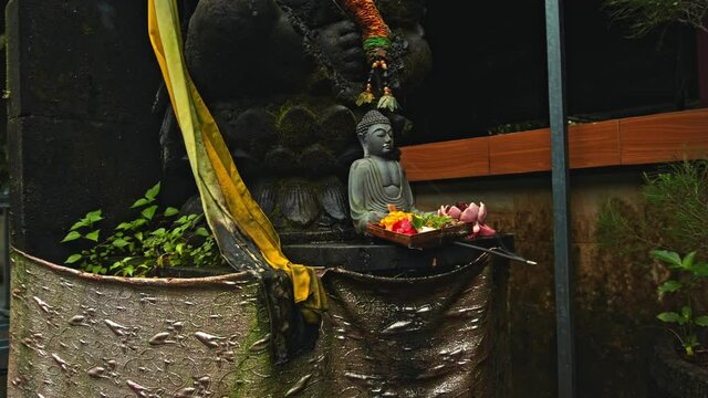 An old stone Budist statue sitting in a lotus pose covered with moss with a wreath of yellow flowers on its neck standing on the street