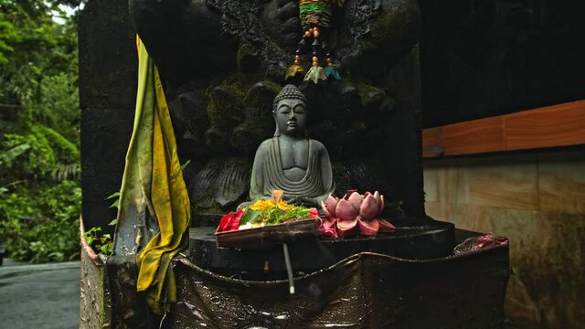 An old stone Budist statue sitting in a lotus pose covered with moss with a wreath of yellow flowers on its neck standing on the street
