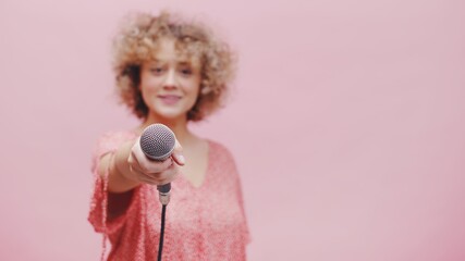 Beautiful young girl holding microphone towards the camera. The focus is on the mic while the girl is blurred in the background. Isolated with pink background studio. Reporter or Journalist concept.  - obrazy, fototapety, plakaty