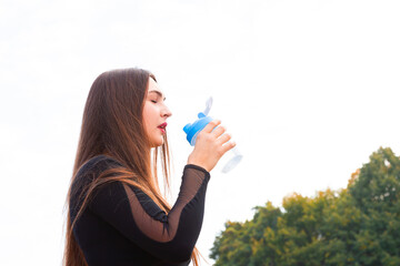 Beautiful young girl drinks water in the park after exercise. Portrait of sporty woman drinking water in park after jogging. Female athlete after training workout. Copy space