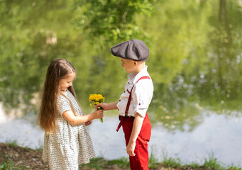 A little boy gives a girl a bouquet of wildflowers. Children stand in the park by the pond.