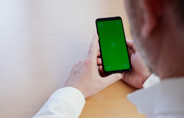 A mock-up image of a hand man in a white shirt holding a black mobile phone with a blank green screen at a wooden table.