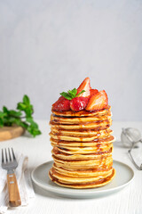 Portion of homemade american organic pancakes or crepes decorated with strawberries and mint leaves served in stack on gray round plate on white wooden table with fork. Vertical image with copy space
