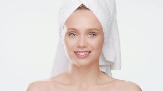 Close-up beauty portrait of cute European young slim woman with towel on her head touches her cheeks after bathing and smiles wide for the camera on white background | Acne prevention concept