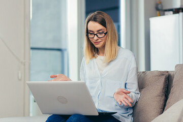 Beautiful blonde woman in casual clothes sitting on a sofa at home, and uses a laptop for video communication with friends and family