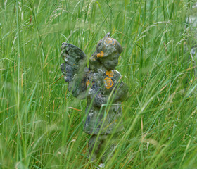 a delightful stone carved angel figurine stands amongt the tall grass in a small English village church graveyard 