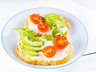 Two toasts with avocado and arugula on a plate. Avocado toasts with cream cheese and cherry tomatoes. Diet food. Close-up