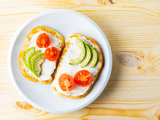 Two toasts with avocado and cherry tomatoes on wooden background. Avocado toasts with cream cheese on a white plate. Copy space. Top view