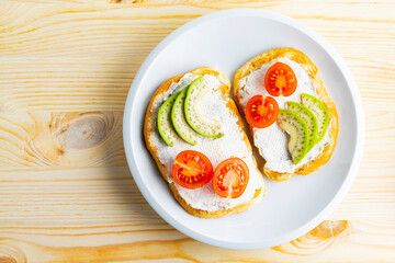 Two toasts with avocado and cherry tomatoes on wooden background. Avocado toasts with cream cheese on a white plate. Copy space. Top view