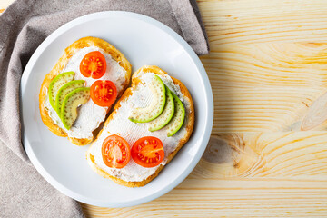 Two toasts with avocado and cherry tomatoes on wooden background. Avocado toasts with cream cheese on a white plate. Copy space. Top view