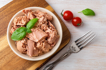 Top view of canned tuna in a bowl, fork and red cherry tomatoes on a white wooden table. Healthy eating snack of preserved tuna meat and fresh vegetables. Low calories tasty seafood. - obrazy, fototapety, plakaty