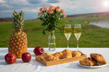 Romantic picnic outdoor. Flowers, glasses of champagne, pineapple, croissants on a tray, bakery and...