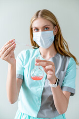 Young woman scientist in medical mask conducts research and makes broad discovery in field of medicine, holding pipette and flat-bottomed flask of red liquid.