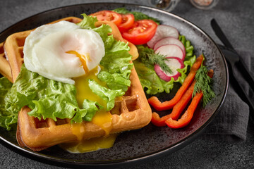 Tasty waffles with poached egg and vegetables on black plate on grey background. Top view.  Table...