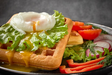 Tasty waffles with poached egg and vegetables on black plate on grey background. Table setting