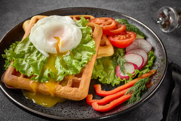 Tasty waffles with poached egg and vegetables on black plate on grey background. Top view.  Table setting
