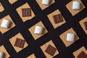 s'mores ingredients. graham cracker squares with chocolate bars, marshmallows on a black background. 