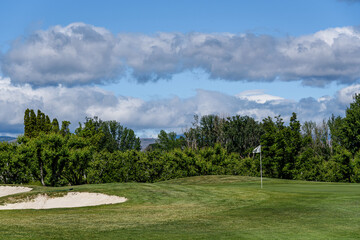 Sunny golf, sand bunker and fairway up to a green with scenic woodland and blue sky in the background
