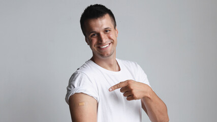 Covid-19 vaccinated caucasian smiling man showing arm with plaster, gray background