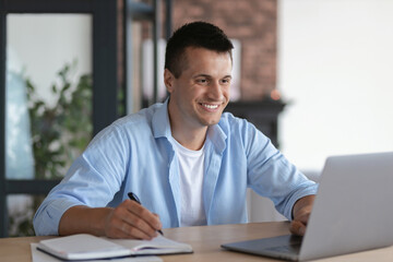 Friendly satisfied caucasian man student, taking notes in notebook during lecture sitting at table in home, online studying at home concept distance education, video call, smiling