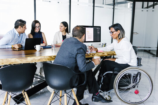 Hispanic transgender businesswoman in wheelchair working on project with group of business people in office, in disability concept and disabled people