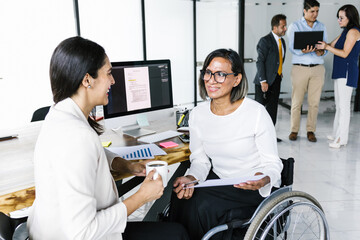 Latin transgender businesswoman in wheelchair and office woman working with computer, in disability...