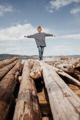 A happy boy in a striped vest walks along the logs on the river bank.