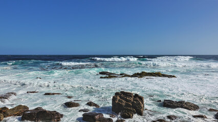 The turquoise waves of the Atlantic Ocean crash against the coastal boulders. Thick white foam on the beach. Clear blue sky. Cape of Good Hope. South Africa