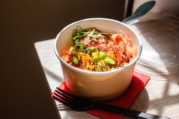 Healthy poke bowl dish with salmon and vegetables in kraft paper packaging on a wooden table. Takeout food. Fitness food. Dinner.